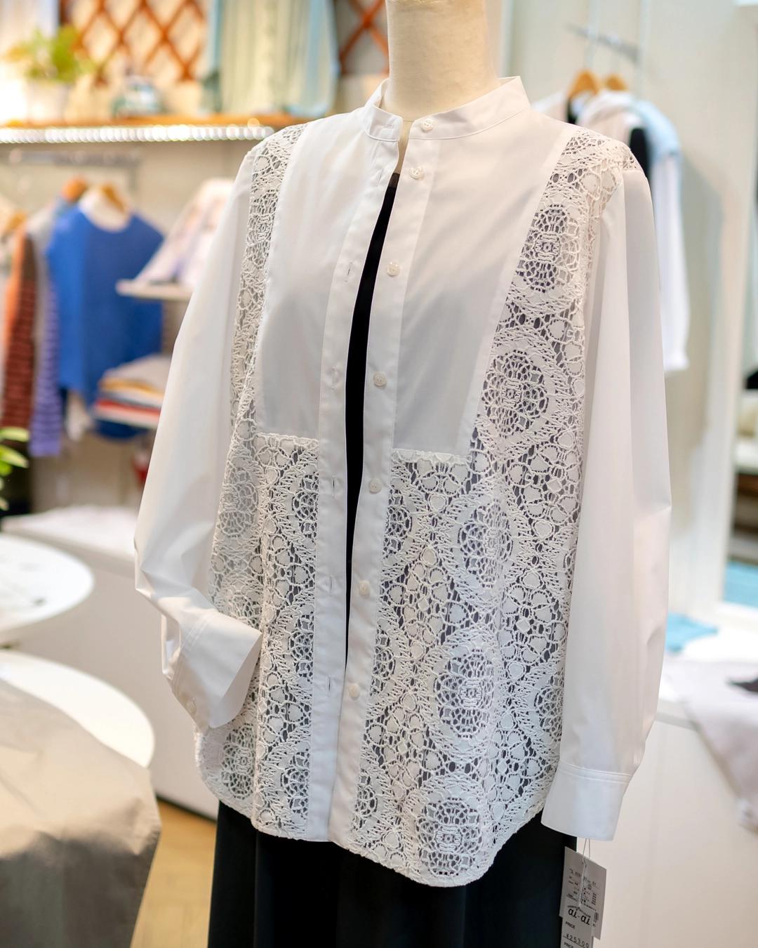 明るい店内に置かれた、白の長袖バンドカラーシャツのディスプレイ。前身頃の左右に、繊細な円形の幾何学模様が連なった大ぶりのレース生地が大胆に切り替えられている。インナーに黒を合わせることで、レースの透け感が際立つスタイリング。清潔感と華やかさを併せ持つデザイン。